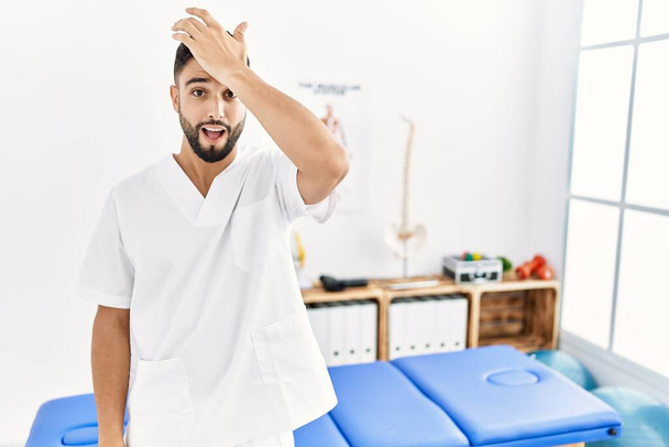 Young handsome man with beard working at pain recovery clinic surprised with hand on head for mistake, remember error. forgot, bad memory concept.  - Photo, Image