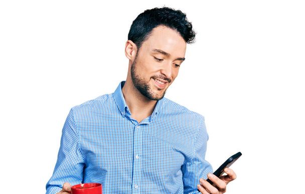 Hispanischer Mann mit Bart, Smartphone und einer Tasse Kaffee, der mit einem fröhlichen und kühlen Lächeln im Gesicht lächelt. Zähne zeigen.  - Foto, Bild
