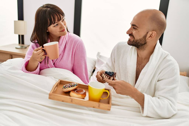 Junges hispanisches Paar frühstückt sitzend auf dem Bett im Schlafzimmer. - Foto, Bild