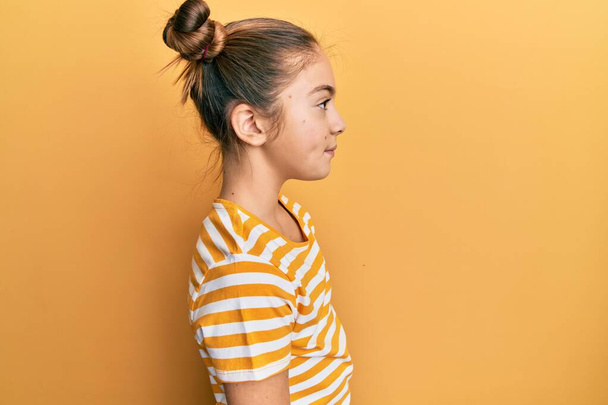 Красивая брюнетка маленькая девочка в повседневной полосатой футболке глядя в сторону, расслабиться профиль позировать с естественным лицом и уверенной улыбкой.  - Фото, изображение