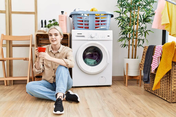 Giovane donna caucasica che beve caffè in attesa di lavatrice in lavanderia - Foto, immagini