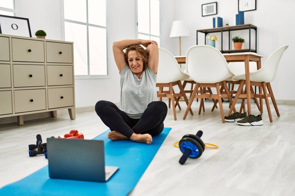 Middelbare leeftijd blanke vrouw glimlachen zelfverzekerd met behulp van laptop stretching thuis - Foto, afbeelding