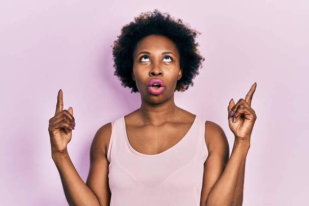 Young african american woman wearing casual sleeveless t shirt amazed and surprised looking up and pointing with fingers and raised arms.  - Foto, Imagem