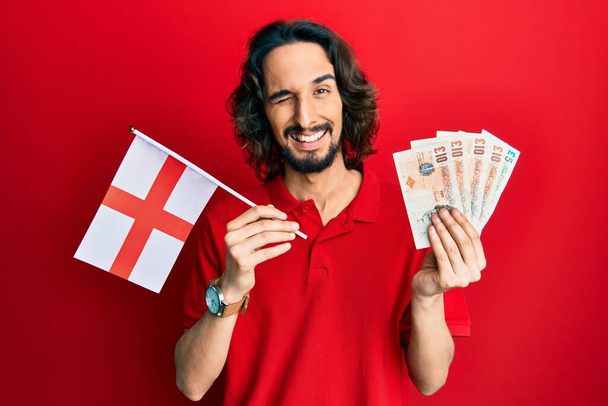 Молодий іспаномовний чоловік тримає прапор Англії і фунти банкнот підморгнув, дивлячись на камеру з сексуальним виразом, веселим і щасливим обличчям
.  - Фото, зображення