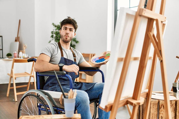 Νεαρός Ισπανός που κάθεται σε αναπηρική καρέκλα ζωγραφίζοντας στο στούντιο τέχνης κάνοντας γκριμάτσες με χείλη, τρελή και κωμική χειρονομία. αστεία έκφραση.  - Φωτογραφία, εικόνα