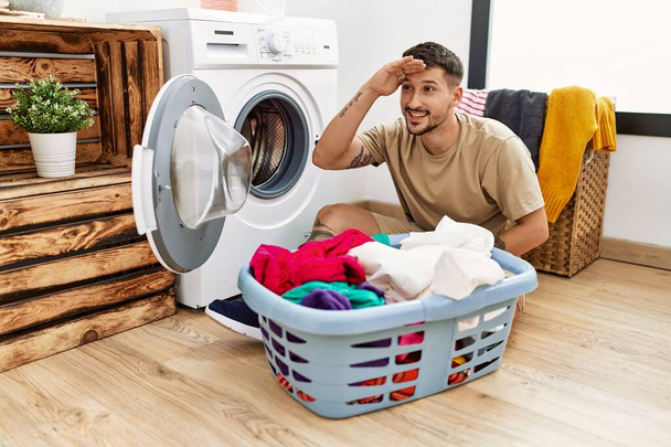 Young handsome man putting dirty laundry into washing machine very happy and smiling looking far away with hand over head. searching concept.  - 写真・画像