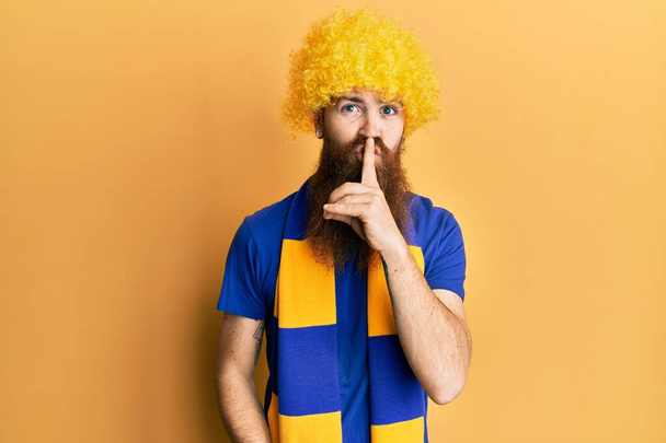 Roodharige man met lange baard voetbal hooligan juichen spel dragen grappige pruik vragen om stil te zijn met de vinger op de lippen. stilte en geheim concept.  - Foto, afbeelding