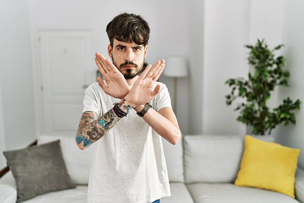 Hispanic man with beard at the living room at home rejection expression crossing arms and palms doing negative sign, angry face  - Фото, зображення