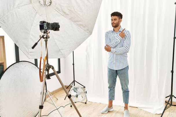 Arab young man posing as model at photography studio pointing with hand finger to the side showing advertisement, serious and calm face  - Foto, Bild