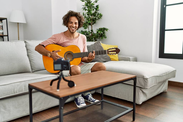 Joven hombre hispano grabando con cámara tocando guitarra clásica en casa. - Foto, imagen