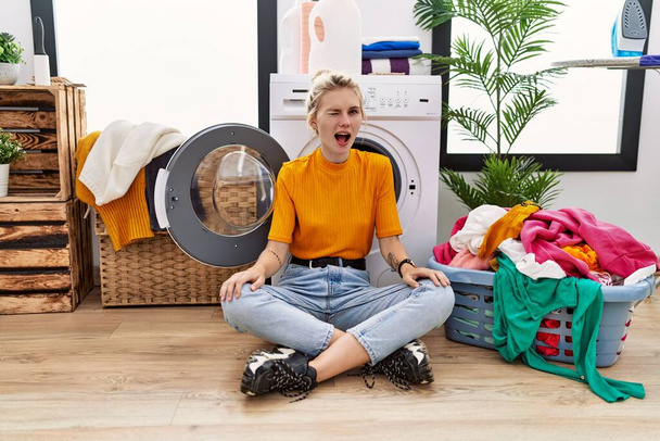 Young blonde woman doing laundry sitting by washing machine winking looking at the camera with sexy expression, cheerful and happy face.  - Φωτογραφία, εικόνα