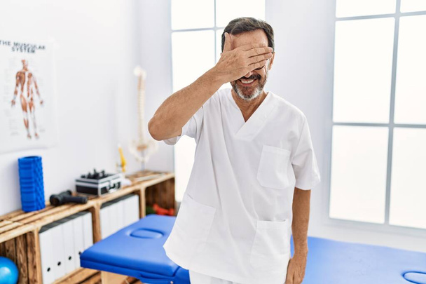 Middle age man with beard working at pain recovery clinic smiling and laughing with hand on face covering eyes for surprise. blind concept.  - Φωτογραφία, εικόνα