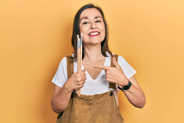 Middle age hispanic woman holding paintbrushes smiling happy pointing with hand and finger  - Photo, Image