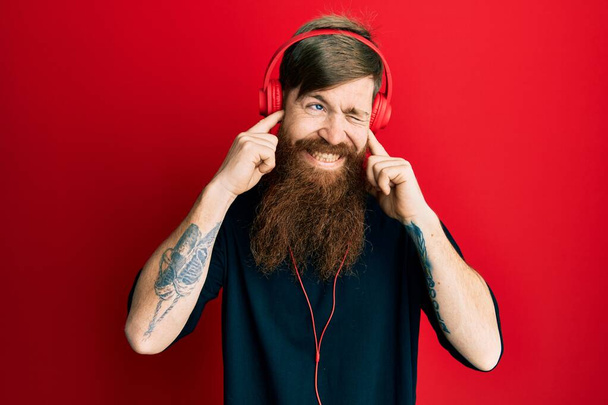 長い髭を生やした赤毛の男は、大きな音楽のノイズに悩まされた表情で指で耳を覆うヘッドフォンを使用して音楽を聞いています。聴覚障害の概念.  - 写真・画像