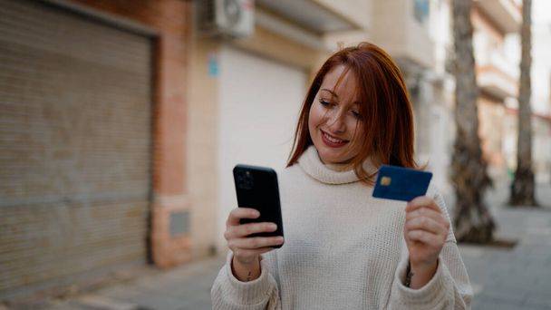 Νεαρή κοκκινομάλλα γυναίκα που χρησιμοποιεί smartphone και πιστωτική κάρτα στο δρόμο - Φωτογραφία, εικόνα