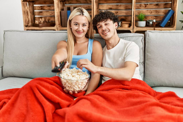 Nuori pari katsomassa elokuvaa ja syömässä popcornia istuu sohvalla kotona. - Valokuva, kuva