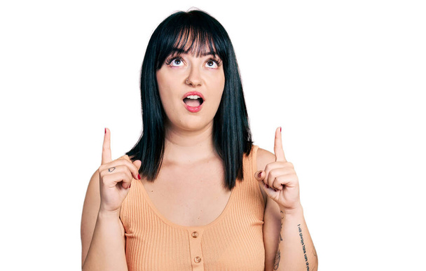 Young hispanic plus size woman wearing casual clothes amazed and surprised looking up and pointing with fingers and raised arms.  - Valokuva, kuva
