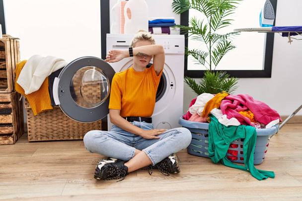 Young blonde woman doing laundry sitting by washing machine covering eyes with arm, looking serious and sad. sightless, hiding and rejection concept  - Photo, Image