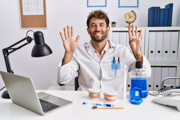 Junger hispanischer Zahnarzt, der in der medizinischen Klinik arbeitet, zeigt mit Finger Nummer neun nach oben und lächelt dabei selbstbewusst und glücklich.  - Foto, Bild