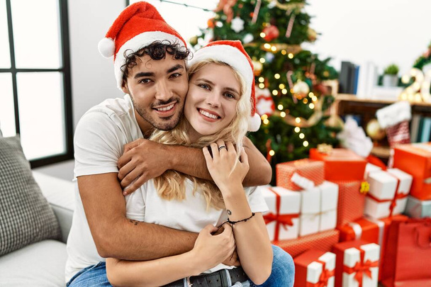 Young couple smiling happy wearing christmas hugging and sitting on the sofa hat at home. - 写真・画像