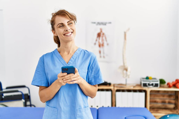 Young caucasian physio therapist girl smiling happy using smartphone at the clinic. - 写真・画像
