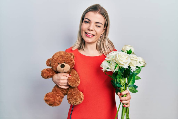 Beautiful caucasian woman holding bear and bouquet of flowers for anniversary winking looking at the camera with sexy expression, cheerful and happy face.  - Foto, Imagem