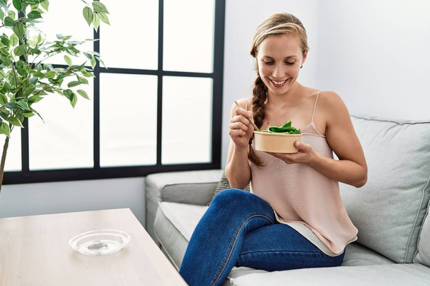 Молодая белая женщина улыбается уверенно поедая салат дома - Фото, изображение