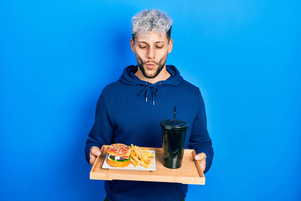 Young hispanic man with modern dyed hair eating a tasty classic burger with fries and soda making fish face with mouth and squinting eyes, crazy and comical.  - Fotó, kép