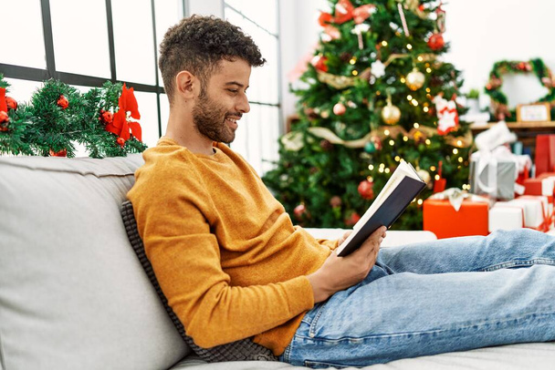 Hombre árabe joven leyendo libro sentado en el sofá por el árbol de Navidad en casa. - Foto, imagen