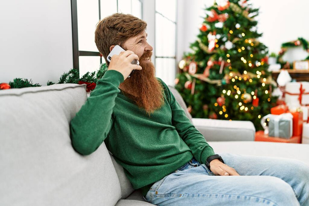 Young redhead man talking on the smartphone sitting by christmas tree at home - 写真・画像