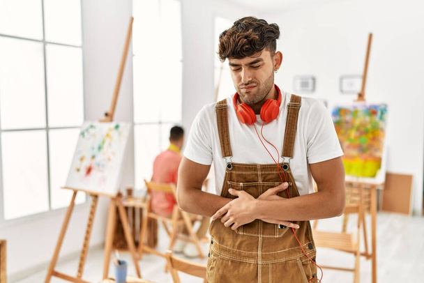 Jonge Spaanse man in de kunststudio met de hand op de maag omdat misselijkheid, pijnlijke ziekte zich onwel voelt. pijnconcept.  - Foto, afbeelding