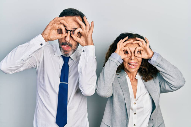 Middle age couple of hispanic woman and man wearing business office uniform doing ok gesture like binoculars sticking tongue out, eyes looking through fingers. crazy expression.  - Foto, Imagem