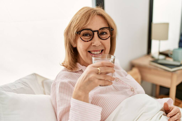 Middelbare leeftijd blonde vrouw glimlachen gelukkig drinken glas water liggend op het bed thuis. - Foto, afbeelding