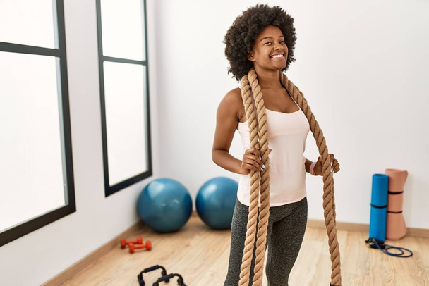 Mladá africká americká žena usmívá sebevědomě drží bojové lano ve sportovním centru - Fotografie, Obrázek
