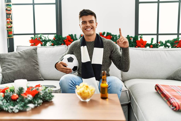 Young hispanic man football hooligan holding ball and beer surprised with an idea or question pointing finger with happy face, number one  - Foto, immagini