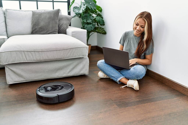 Young caucasian girl controlling robot vacuum cleaner with laptop sitting on the floor at home. - Φωτογραφία, εικόνα