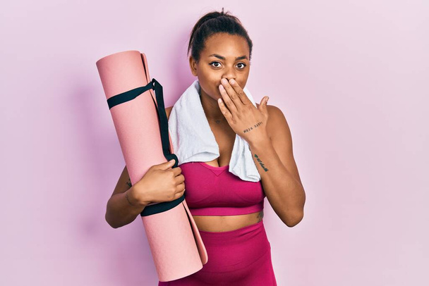 Joven afroamericana americana con ropa deportiva sosteniendo esterilla de yoga cubriendo la boca con la mano, conmocionada y temerosa de equivocarse. expresión sorprendida  - Foto, imagen