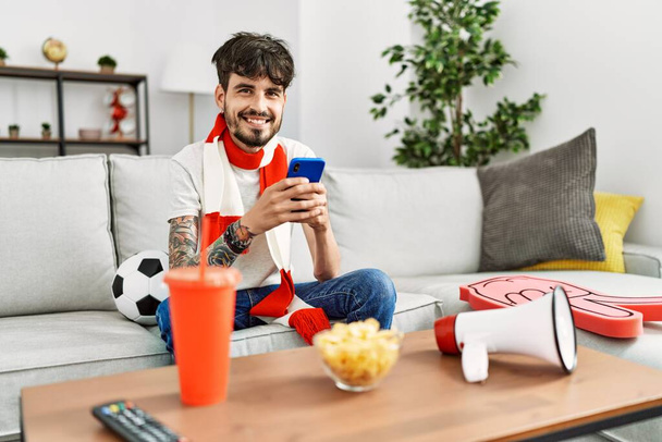 Young hispanic hooligan man using smartphone supporting soccer team at home - Photo, Image