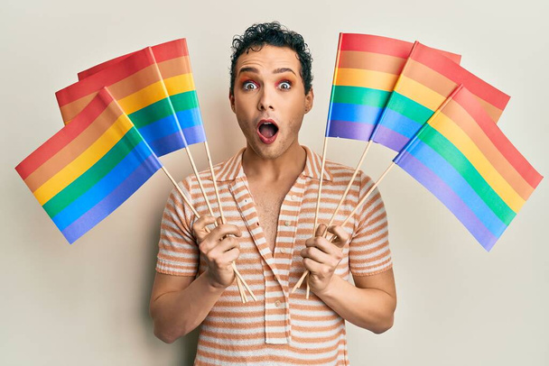 Handsome man wearing make up holding rainbow lgbtq flags afraid and shocked with surprise and amazed expression, fear and excited face.  - Zdjęcie, obraz