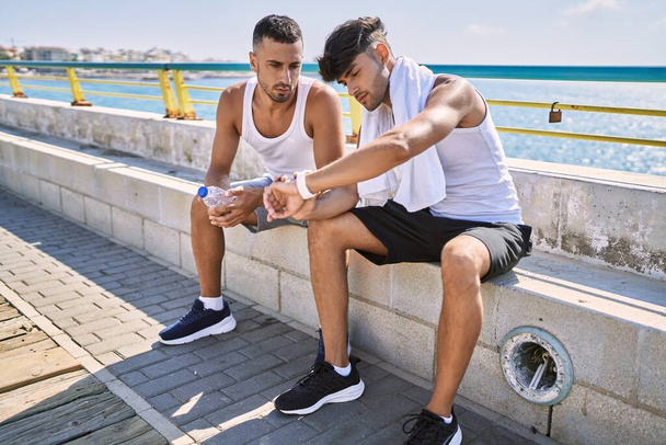 Two hispanic men sporty couple smiling confident looking watch sitting on bench at seaside - Fotoğraf, Görsel