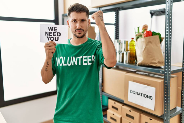 Young hispanic man wearing volunteer t shirt holding we need you banner annoyed and frustrated shouting with anger, yelling crazy with anger and hand raised  - Fotoğraf, Görsel