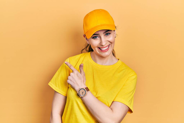 Young hispanic woman wearing delivery uniform and cap cheerful with a smile of face pointing with hand and finger up to the side with happy and natural expression on face  - Photo, Image