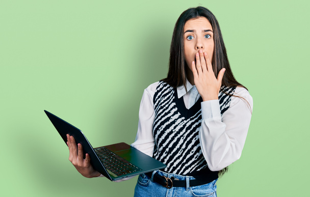 Young brunette teenager working using computer laptop covering mouth with hand, shocked and afraid for mistake. surprised expression  - Foto, Bild