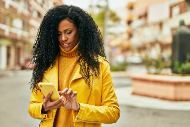 アフリカ系アメリカ人の中年女性が市内のスマートフォンを使って笑顔で幸せ. - 写真・画像