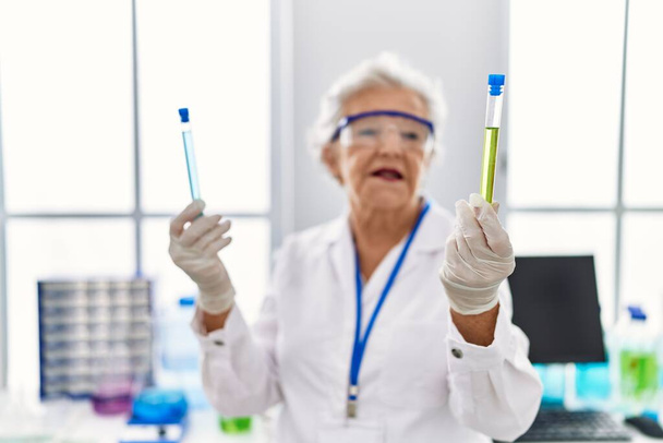Mujer mayor de cabello gris con uniforme científico sosteniendo tubos de ensayo en el laboratorio - Foto, imagen