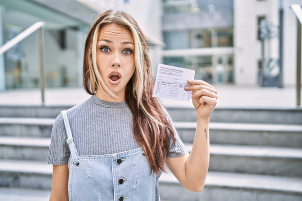Joven mujer caucásica sosteniendo tarjeta de registro covid asustado y sorprendido con la boca abierta para sorpresa, cara de incredulidad  - Foto, imagen