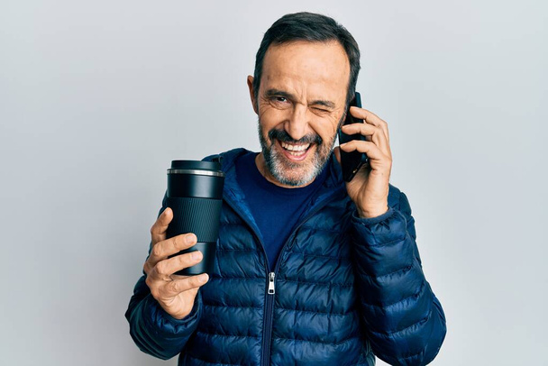 Middle age hispanic man talking on the smartphone and drinking a cup of coffee winking looking at the camera with sexy expression, cheerful and happy face.  - Foto, Imagem