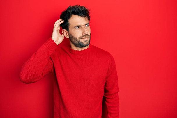 Handsome man with beard wearing casual red sweater confuse and wondering about question. uncertain with doubt, thinking with hand on head. pensive concept.  - Photo, Image