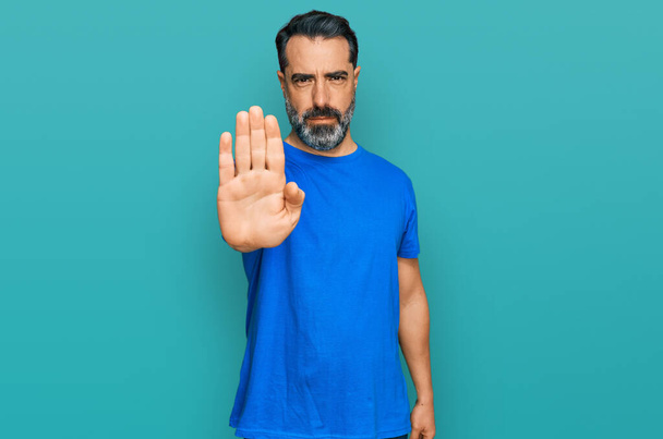 Hombre de mediana edad con barba con camiseta azul casual haciendo parar de cantar con la palma de la mano. expresión de advertencia con gesto negativo y grave en la cara.  - Foto, Imagen