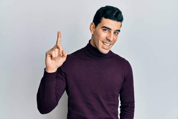 Un bel homme hispanique portant un pull à col roulé souriant avec une idée ou une question pointant du doigt avec un visage heureux, numéro un  - Photo, image